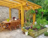 Une pergola en bois pour fabriquer une pergola pour plante grimpante, avec table et chaises, et un parterre de fleurs.