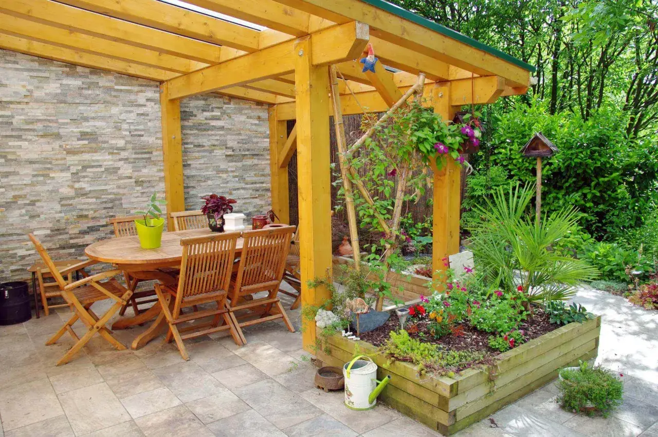 Une pergola en bois pour fabriquer une pergola pour plante grimpante, avec table et chaises, et un parterre de fleurs.