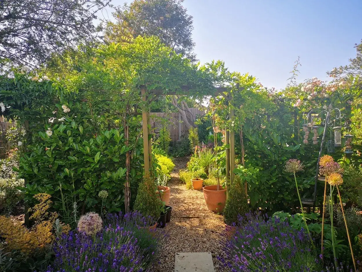 Un chemin de gravier mène à travers une arche de jardin en bois, ornée de roses et de verdure luxuriante.