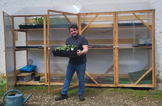 Un homme souriant tient un plateau de jeunes plants devant une serre facile à fabriquer, remplie d'étagères de semis.