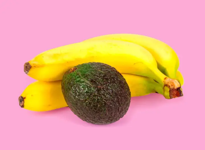 Un avocat vert foncé repose à côté d'une grappe de bananes jaunes mûres. La banane est connue pour aider à faire murir l'avocat.